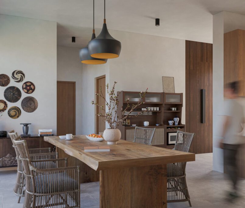 Une cuisine moderne et une salle à manger avec des meubles en bois, des chaises en osier et un décor minimaliste. Des œuvres d'art murales sont accrochées à côté des armoires et une personne floue marche près d'un plan de travail équipé d'appareils de cuisine. Les tons neutres créent une atmosphère chaleureuse.